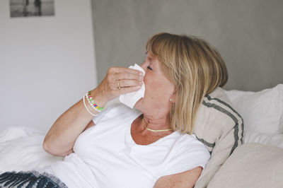 Sick senior woman blowing nose while lying on bed at home