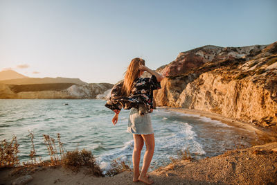 Back view anonymous fit female in mini skirt standing on rough rocky seashore and touching long hair under clear blue sky in fyriplaka milos