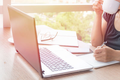 Businesswoman drinking coffee while writing on book by laptop in office