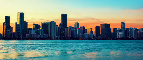 Sea by modern buildings against sky during sunset