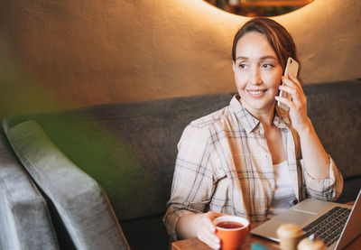 Adult charming brunette woman in plaid shirt working with laptop using mobile phone at cafe