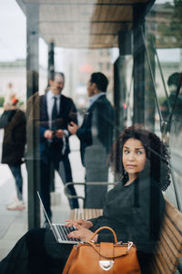 Side view of mid adult businesswoman looking away while sitting with laptop at bus shelter seen from glass