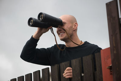 Close-up of mid adult man looking through binoculars while standing against sky