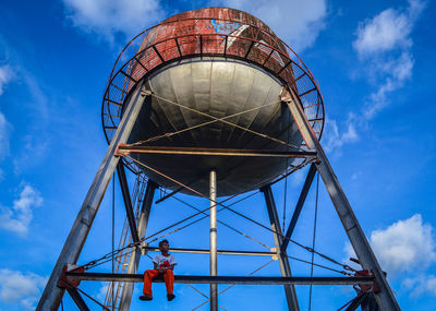 Low angle view of man sitting on water tower against blue sky
