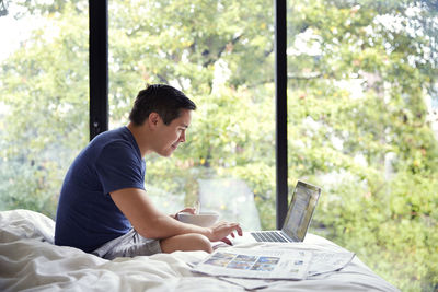 Side view of man using laptop while having breakfast on bed by window