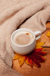 Coffee with milk foam in a white mug, autumn red and yellow maple leaves and a knitted beige sweater