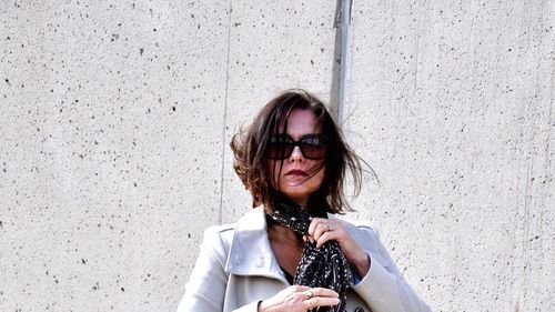 Portrait of young woman tying scarf while standing against wall