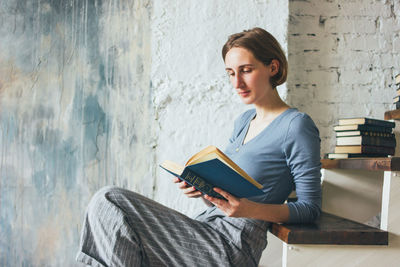 Young woman reading book while sitting on steps against wall