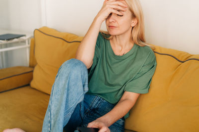 Young sad woman sitting on the couch holds her hand on her sore head. person