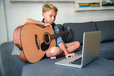 Full length of boy using laptop while learning guitar on sofa at home