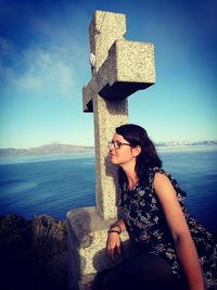 Young woman looking away while leaning by cross against sea