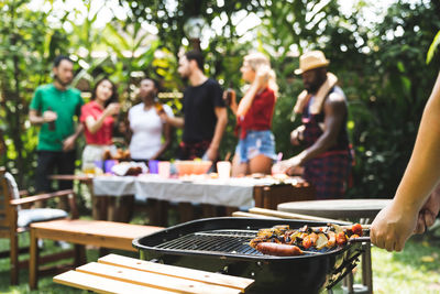 Midsection of woman preparing meat on barbecue against friends enjoying in garden during party