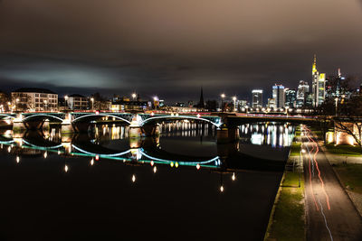 Illuminated bridge over river by buildings against sky at night