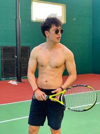 Shirtless young man holding tennis racket while standing on court