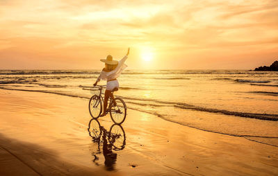 Rear view of man riding bicycle on beach against sky during sunset
