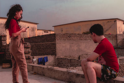 Woman using mobile phone with friend on terrace against sky during sunset