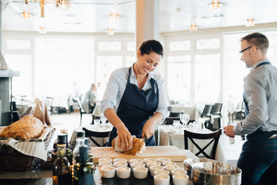 Smiling owner cutting bread on table while coworker in background