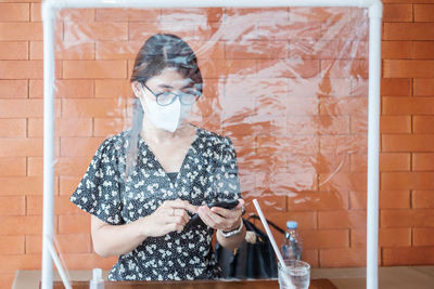 Portrait of young woman using phone while standing against brick wall