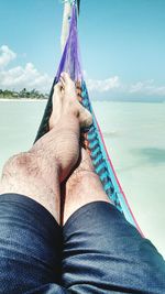 Low section of man relaxing in hammock over sea against sky