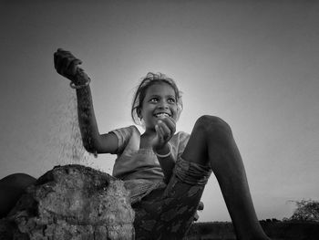 Smiling girl looking away while putting sand on rock against clear sky