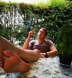 Young man smoking cigarette while sitting on table against plants 