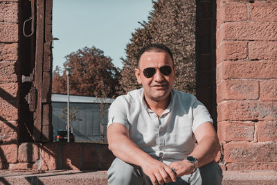 Portrait of man wearing sunglasses sitting against brick wall