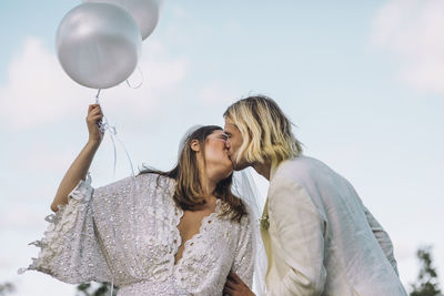 Bride holding white helium balloons kissing groom against sky