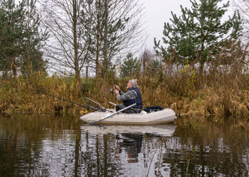 Fisherman in a white boat, river bank with bare trees and bushes, shore reflection in river water