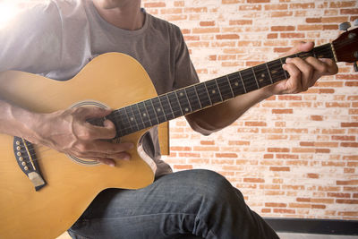 Midsection of man playing guitar while sitting against brick wall