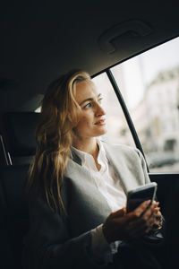Confident female entrepreneur with smart phone looking through window while sitting in taxi
