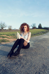 Portrait of young woman sitting on road against clear sky during sunset