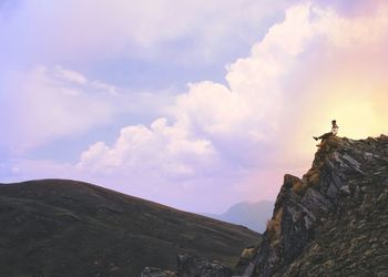 Mid distant view of man sitting on top of mountain against cloudy sky