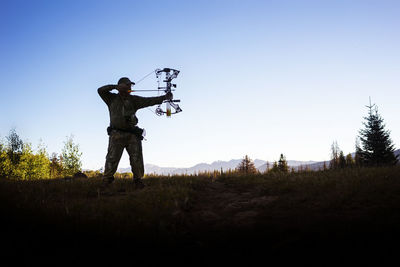 Hunter aiming with bow and arrow while standing against clear sky