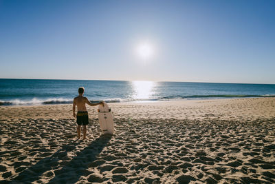 Rear view of man standing with surfboard on beach against clear sky