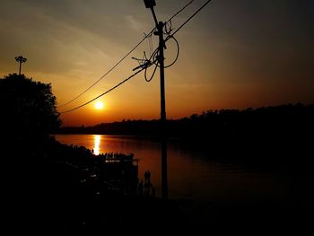 Silhouette electricity pylon by lake against sky during sunset