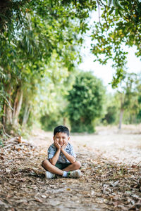 Portrait of cute boy with hands on chin sitting against trees