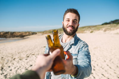 Smiling young man toasting alcoholic drink bottle with friend at beach during sunny day