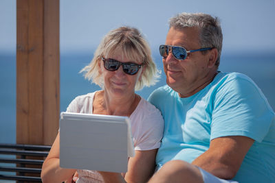 Mature couple wearing sunglasses while using digital tablet at beach