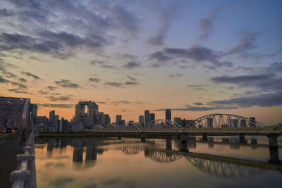 Bridge over river by buildings against sky during sunset