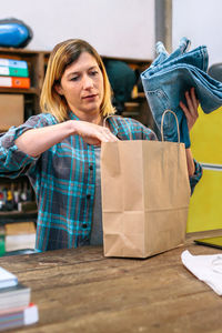 Concentrated woman shop assistant putting jeans into a ecologic paper bag to online shop customer