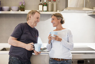 Cheerful senior couple talking while holding coffee mugs in kitchen