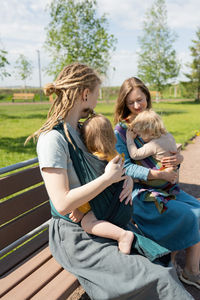 Happy mothers with their babies sitting on bench at park on sunny day