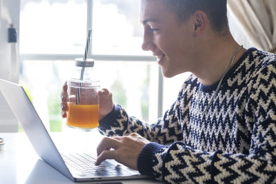 Smiling teenager boy holding juice while using laptop at home