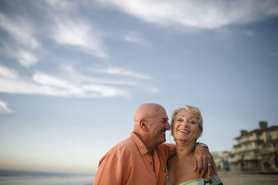Smiling senior couple standing at beach against sky during sunset
