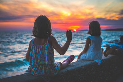 Girls sitting on retaining wall against sky during sunset