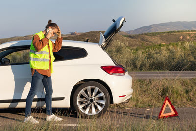 Anxious and desperate young man talking on the phone asking for help with his broken car behind him