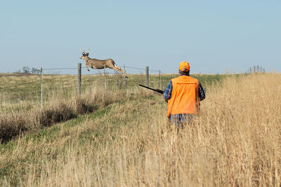 Rear view of man with gun while deer jumping over fence on field against sky