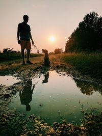 Rear view of man standing by puddle against sky during sunset