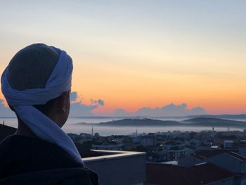 Rear view of man looking at cityscape against sky during sunset