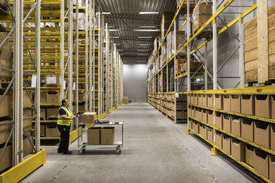 Full length side view of mature female warehouse worker pushing cart on aisle in industrial building